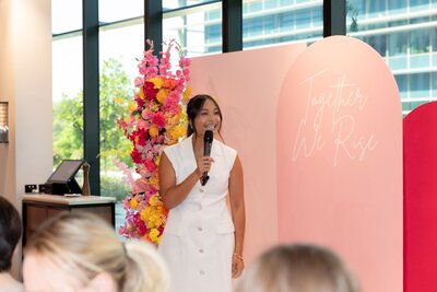 Trang Nova speaking at a women's conference wearing a white dress standing in front of a pink and yellow floral installation. 