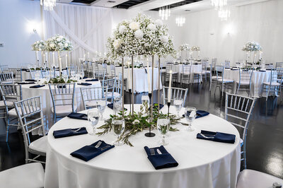 A round reception table at SoHo63, a Chandler wedding and event venue, is set with navy napkins, glassware, and a tall centerpiece of white flowers and greenery. The surrounding space features silver chiavari chairs, soft drapery, and modern chandeliers for a clean, classic look.