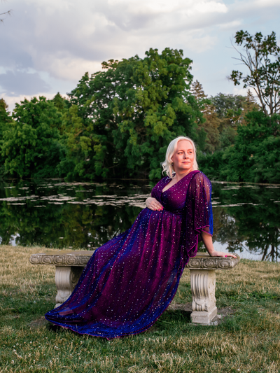 A blonde pregnant woman sits on a bench in front of a lake, looking toward the sunset.