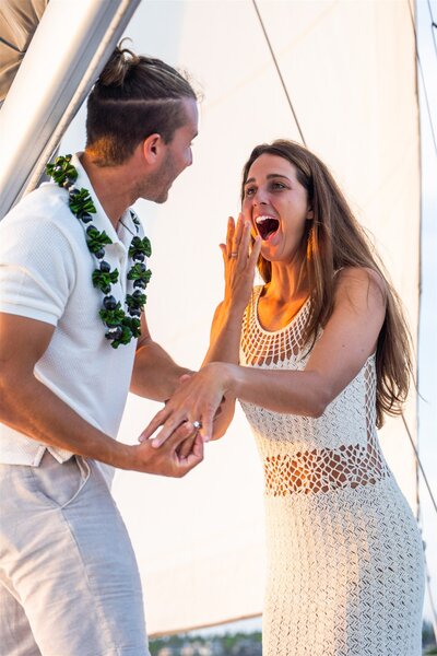 Woman reacting with excitement as her partner holds her hand during a sailboat proposal in Hawaii, smiling and showing her engagement ring.