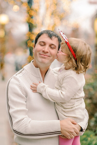 Young daughter in a white dress and red reindeer antlers kissing Dad's cheek during their Christmas photos in Chicago, IL