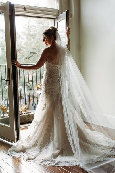 Bride looks out over St. George Street from the balcony doors of The Plaza Suite, wrapped in soft morning light and lace veil.