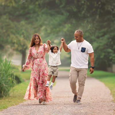 Family of three walking together on a tree-lined path during a summer outdoor session, with their young child smiling and running ahead.