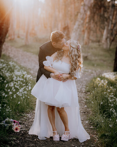 Bride in a short dress being cuddled from behind by groom amongst white wildflowers