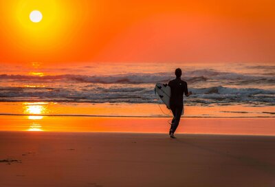 A surfer walking barefoot along a sandy beach at sunset, holding a surfboard under his arm as the waves glisten in the warm orange glow. This picturesque coastal moment captures the adventure and serenity offered through vacation fundraising for charities, inspiring both relaxation and support for important initiatives.