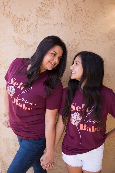 mom and daughter wearing self-love babe women's t-shirts and holding hands while looking at each other