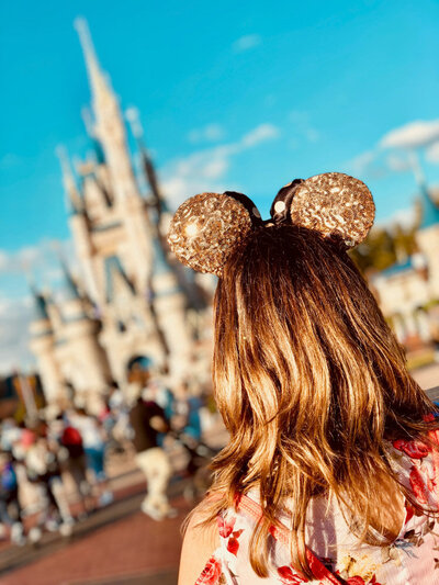 A woman with long hair wearing gold glitter mouse ears stands with her back to the camera while Cinderella's Castle  at Walt Disney World stands in the image background.