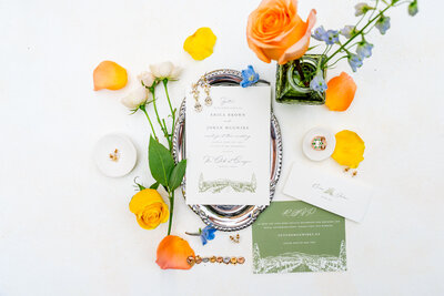 A colorful wedding day flat-lay for Kassie and Alex’s wedding at The Club at Corazón in Dublin, Ohio. The arrangement features their invitation suite with green and white details, gold jewelry, floral accents in yellow, orange, and blue, and a silver tray on a white background.