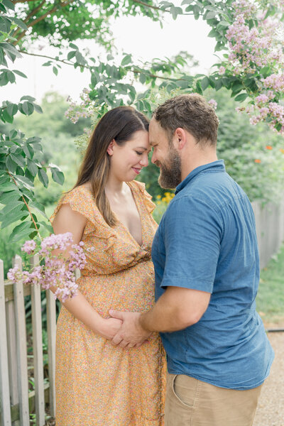 A couple expecting their first child holds hands and smiles at each other, standing under a flowering tree.