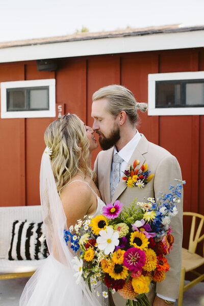 Bride and groom kiss at Ojai Rancho Inn/ The bride is holding a colorful wildflowers bouquet and the groom has a floral pocket square.