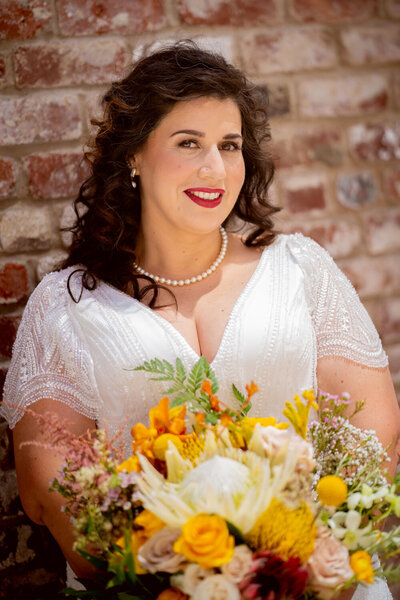 Smiling bride holding a colorful bouquet, posing by a brick wall