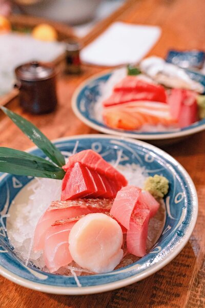 Two bowls of fresh sashimi featuring a variety of fish, including tuna and salmon, are artistically arranged on ice. The dishes are garnished with green leaves and wasabi, served in blue patterned bowls on a wooden table.