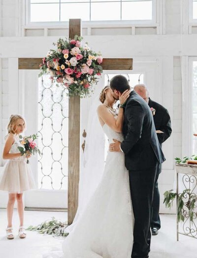 A bride and groom kissing on wedding day in front of a cross covered in florals