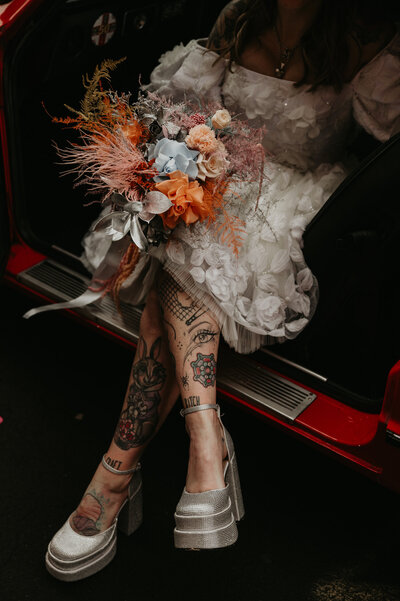 A bride sits in a vintage sports car with her legs crossed and wedding bouquet on her lap. Her dress is short so you can see her tattooed legs. 