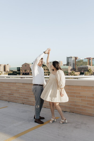 Guy holding hands spinning girl wearing short white poof dress at golden hour during rooftop proposal in Phoenix, Arizona.