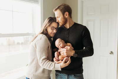 A cozy family moment captured indoors, with a mother and father cradling their newborn baby near a softly lit window. The father gently kisses the mother’s head while she looks down lovingly at their sleeping baby. The natural light enhances the warmth and tenderness of the scene.