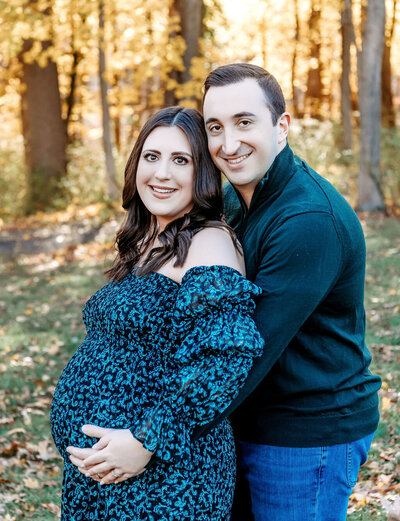 Fall maternity portrait by Stamford CT photographer showing expecting couple in coordinated teal and patterned dress among autumn foliage