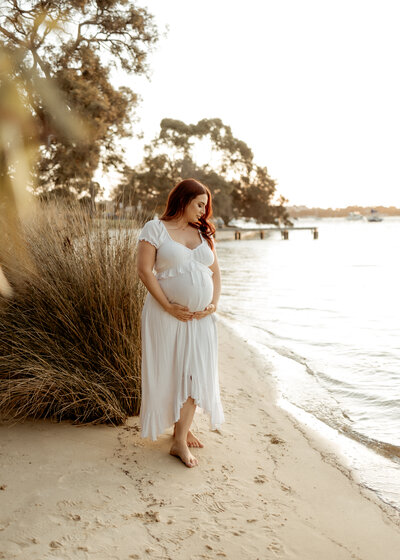 A serene maternity photo taken on a sandy shoreline in Perth, featuring a glowing pregnant woman in a flowing white dress, gently cradling her belly as the golden light of sunset reflects off the water.