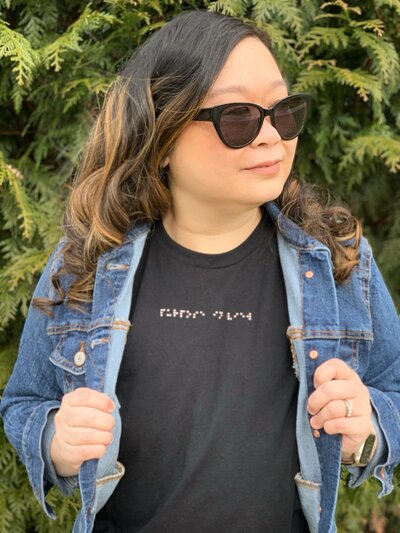 Anne, an Asian woman, is looking off to her left shoulder wearing sunglasses and a black Aille Design T-Shirt with Braille words "purpose in view". She is wearing a denim jacket on top.
