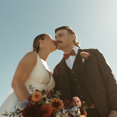 A bride gently kisses the groom’s cheek in an outdoor wedding portrait, glowing in warm sunlight.