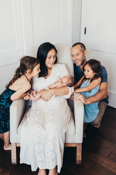 a mother holds her newborn baby while dad and two sisters huddle around the mom's chair during their newborn photoshoot in their encinitas home