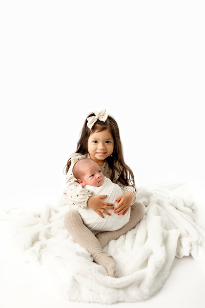 Big sister smiling holding newborn baby brother in her lap, neutr5al colors, white background, clovis newborn photograpehr