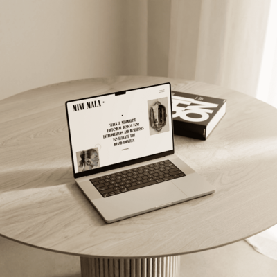 A laptop on a round table displays the MINI MALA website, showcasing a sleek and minimalist design with the tagline "Sleek & Minimalist Editorial Design for Entrepreneurs and Businesses to Elevate the Brand Identity."