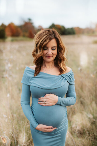 Pregnant mom wearing a light blue dress looking down and cupping belly at Stoney Creek Metro park