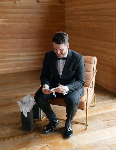 A man in a tuxedo sits in a modern chair, smiling while reading a note. A gift bag with tissue paper is on the wooden floor, creating a warm, intimate atmosphere.