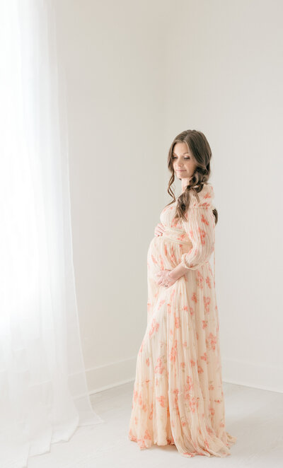 Pregnant woman standing next to window in an all white studio in New Jersey