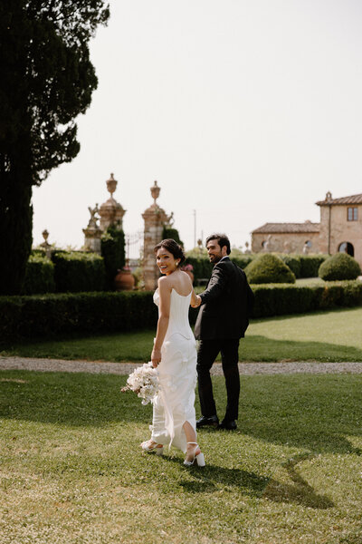 A newlywed couple holding hands and walking as they look over their shoulders 