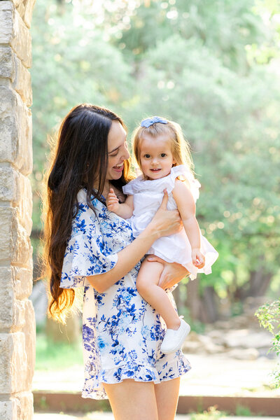 A mother stands under an arch at Mayfield Park. She holds her daughter in her arm, dress in a white dress with a blue bow in her hair. The little girl smiles at the camera while the mother looks at her smiling, in this sweet shot captured by an Austin Family Photographer