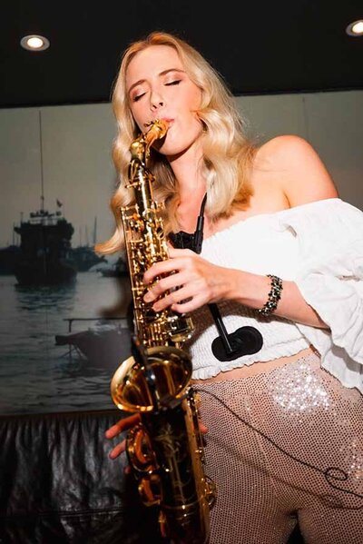 Blonde female saxophonist playing live at an indoor event wearing a white off-shoulder top and sequin pants.