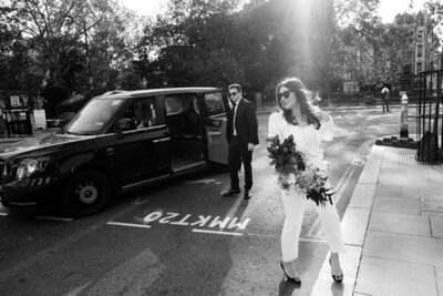 cinematic candid wedding photo of bride in white suit and groom in black tie exiting a taxi in london 