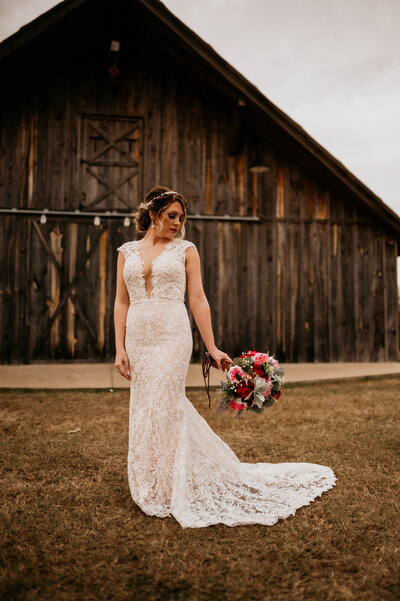 Arkansas wedding photographer captures bride in a lace wedding dress as she holds her burgundy floral bouquet as she stands in front of an old wooden barn