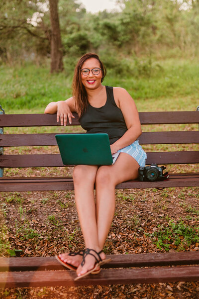 A Texas-based branding photographer sits on a bench with a laptop and camera during a personal branding session in Challenger 7 Park (Houston), surrounded by greenery.
