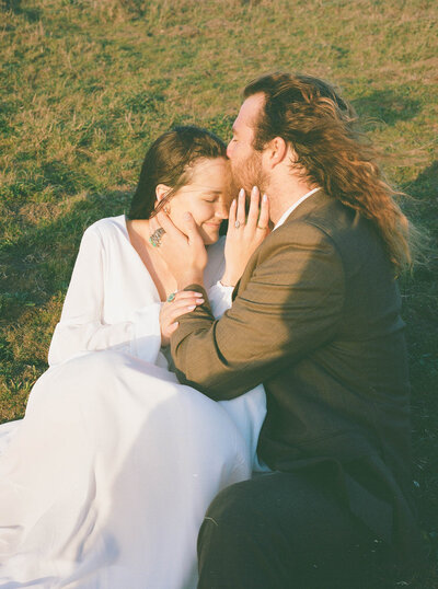 Couple in wedding attire sitting on grass cuddling in while on partner kisses the other's forehead