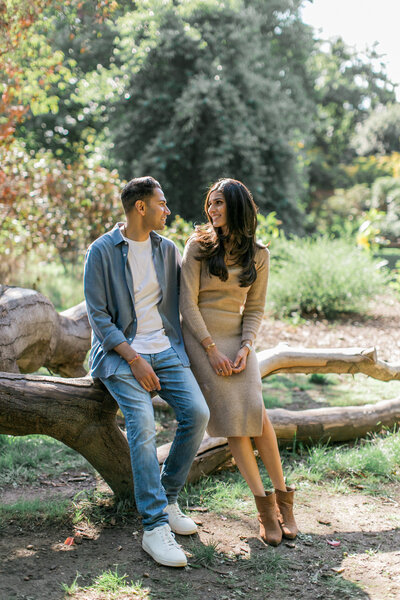 An engaged couple sitting on a fallen tree, and smiling at each other.