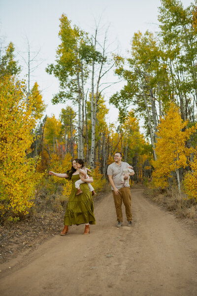 Mom and dad are snuggled together standing up as their boy runs around them in a field with snowy mountains behind them