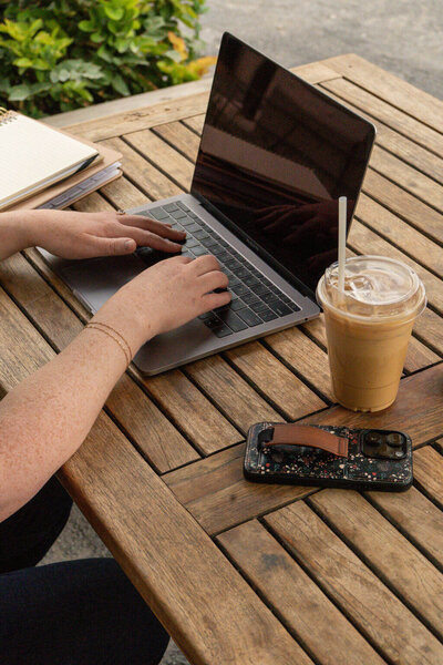 Virtually Robyn, a virtual assistant, is typing on a laptop at a wooden outdoor table, with an iced coffee, a smartphone, and a notebook nearby. Green plants are visible in the background.