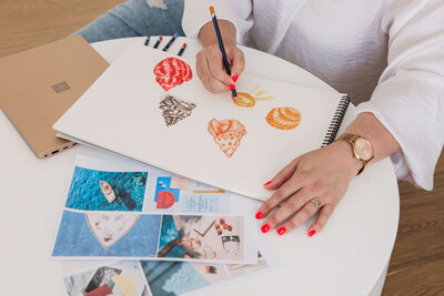 Close up of Leith drawing with laptop and moodboard images scattered around, bright red nail polish