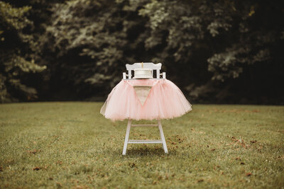 Picture of a white high chair with a white cake on top of it with the number 1 and a pink tutu skirt around high chair top with a banner with the number 1.