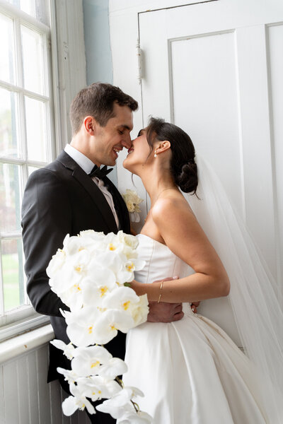 a newly married couple are taking their portraits. They are leaning in for a kiss while she holds a flower bouquet. Both are dressed in suits and wedding dress. 