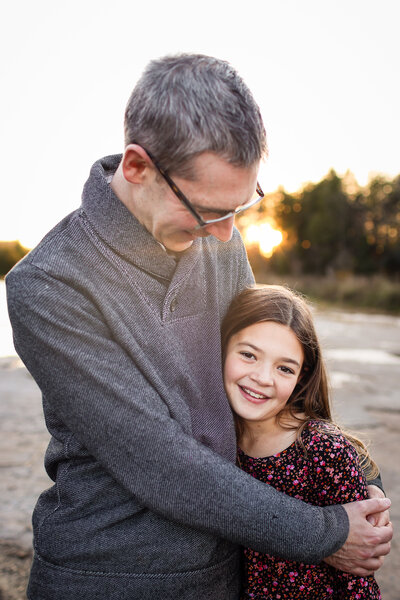 Tender moment between a father and his young daughter, with the father gently hugging her. The girl looks confidently and warmly at the camera in her pink flowered dress. The sun sets behind them.