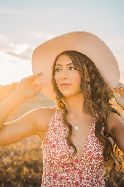 Senior girl with long dark wavy hair wearing a cream wide brimmed hat and a pink flower dress during sunset