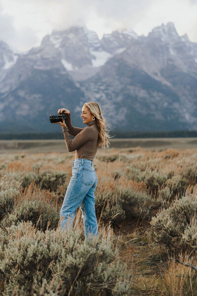 woman standing in a field smiling while holding a camera with mountains in the background