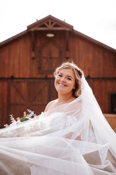 Bride smiling with veil flowing in front of rustic barn