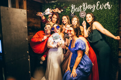 a bride and 8 wedding guests in  a photo booth holding and wearing various props
