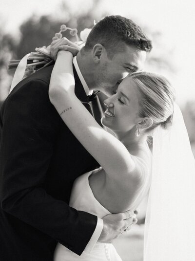 Timeless black-and-white wedding photo of groom kissing bride’s forehead, capturing genuine love and connection.