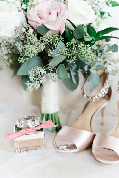 Bridal details in blush and cream, including beige shoes, blush perfume in glass bottle, and a white and cream bouquet